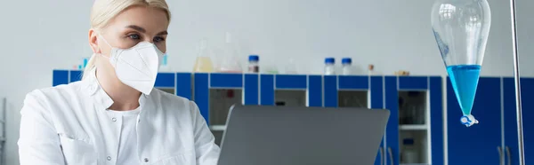 Blonde scientist in protective mask and white coat using laptop near flask with liquid in lab, banner — Fotografia de Stock