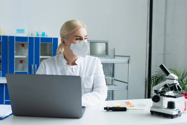 Scientist in protective mask using laptop and looking at microscope in lab — Stock Photo