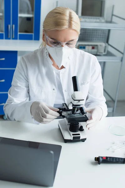 Ученый в защитной маске работает с микроскопом возле ноутбука в лаборатории — стоковое фото