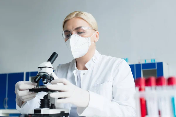 Scientist in goggles and latex gloves using microscope near blurred test tubes in lab — стоковое фото