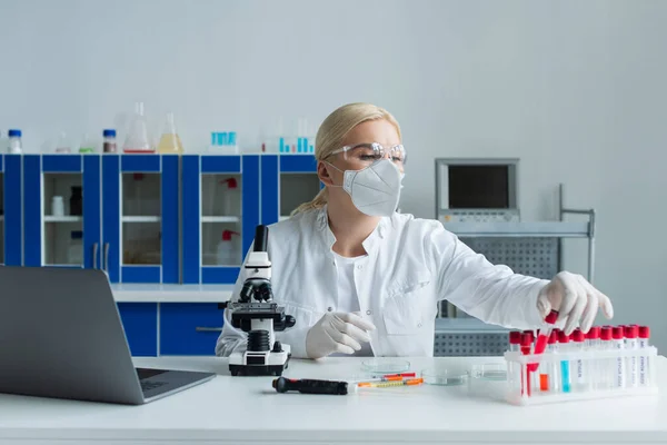 Cientista em máscara protetora levando tubo de teste perto de laptop e microscópio em laboratório — Fotografia de Stock