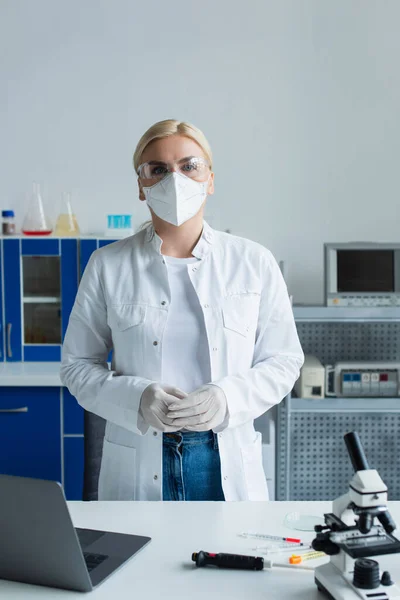 Scientist in goggles and protective mask standing near microscope and laptop in lab — Foto stock