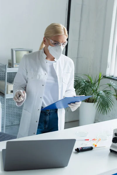 Blonde scientist in goggles holding clipboard near laptop and syringes in lab - foto de stock