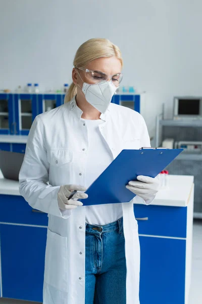Cientista loira em máscara protetora segurando prancheta em laboratório — Fotografia de Stock