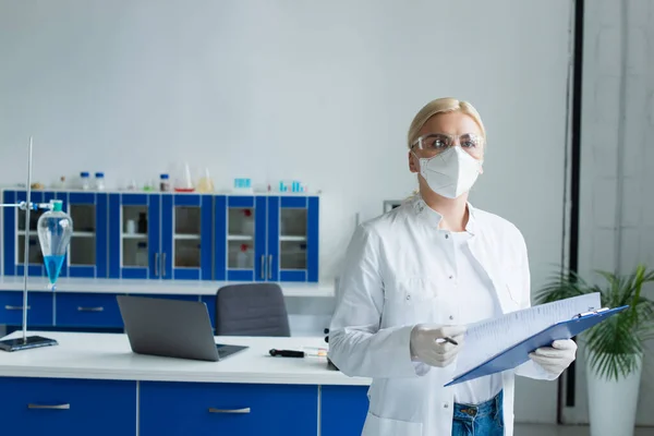 Scientist in googles and protective mask holding clipboard and looking at camera in lab — Stock Photo