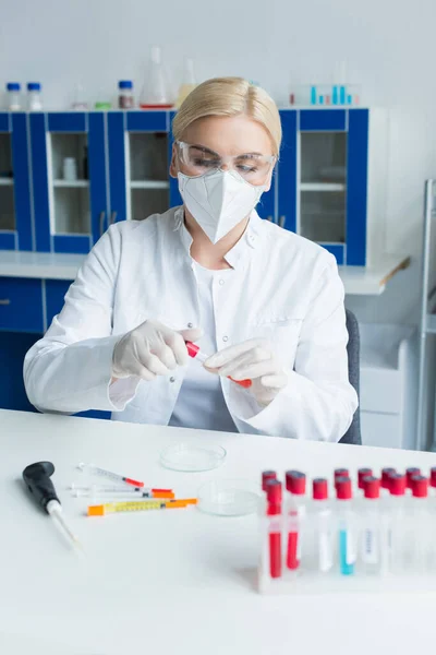 Blonde scientist in protective mask holding test tube near syringes in laboratory - foto de stock