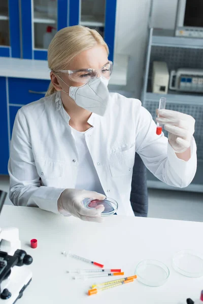 Cientista em óculos e máscara segurando tubo de ensaio e placa de Petri perto de seringas em laboratório — Fotografia de Stock