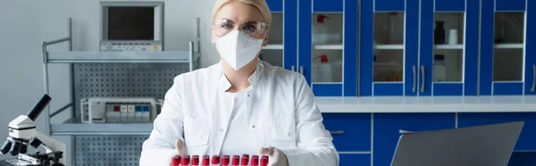 Scientist in goggles looking at camera near test tubes and microscope in lab, banner — Stock Photo
