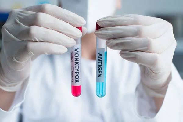 Ausgeschnittene Ansicht eines verschwommenen Wissenschaftlers in Latexhandschuhen, der Reagenzgläser mit Antigen und Affenpocken-Schriftzug im Labor hält — Stockfoto