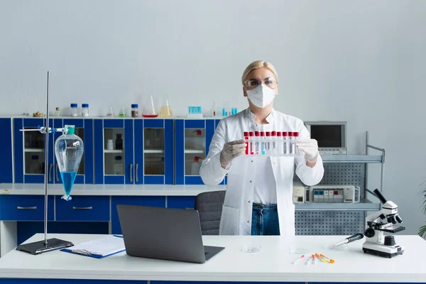 Wissenschaftler mit Schutzbrille hält Reagenzgläser mit Affenpocken-Schriftzug in der Nähe von Laptop und Kolben im Labor — Stockfoto