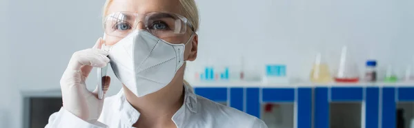 Вчений в окулярах і захисній масці говорить на мобільному телефоні в лабораторії, банер — стокове фото