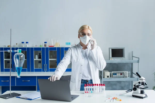 Scientist in goggles and mask talking on smartphone near test tubes and microscope in lab - foto de stock