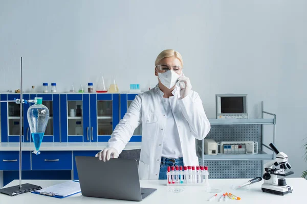 Scientist in goggles and mask talking on smartphone near test tubes and microscope in lab — Stock Photo