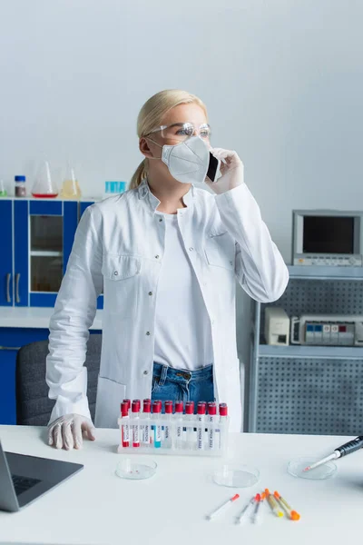 Researcher in medical mask talking on smartphone near test tubes and laptop in lab - foto de stock