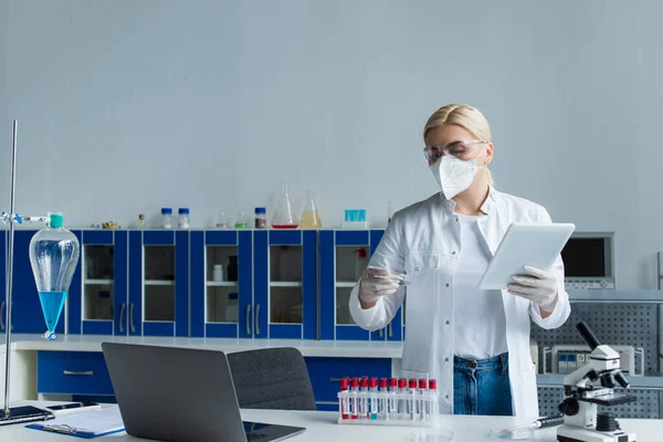 Scientist in protective mask holding petri dish and digital tablet in laboratory — Photo de stock