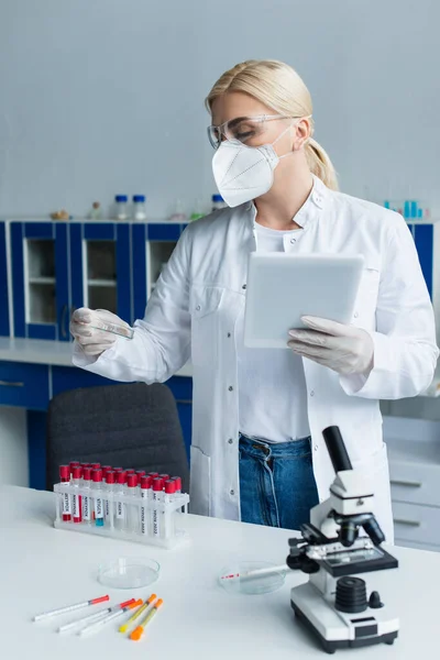 Scientist in protective mask and goggles holding petri dishes and digital tablet near test tubes in lab — Stock Photo