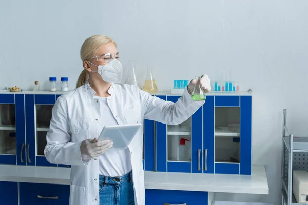 Researcher in protective mask holding flask and digital tablet in laboratory — Stock Photo