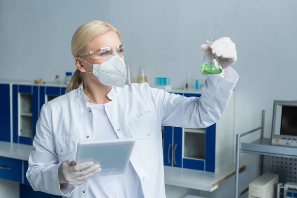 Scientist in protective mask holding flask with liquid and digital tablet in laboratory — Stock Photo
