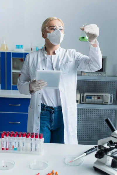 Cientista em máscara protetora segurando frasco e tablet digital perto de tubos de ensaio em laboratório — Fotografia de Stock