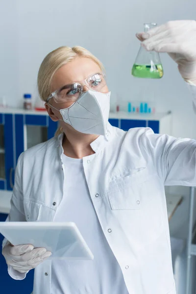Ученый в очках смотрит на размытую фляжку и держит цифровой планшет в лаборатории — стоковое фото