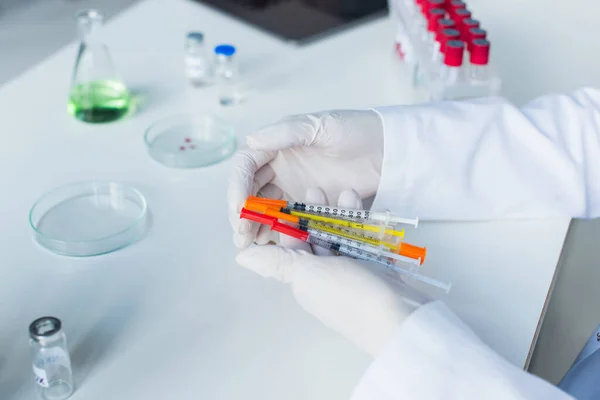 Обрезанный вид ученого, держащего шприцы рядом с вакциной и чашками Петри в лаборатории — стоковое фото