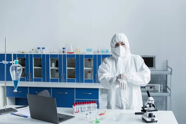 Ученый в защитном костюме и латексных перчатках стоит рядом с пробирками и микроскопом в лаборатории — стоковое фото