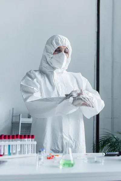 Вчений в захисному костюмі і масці, що стоїть біля розмитих пробірки в лабораторії — стокове фото