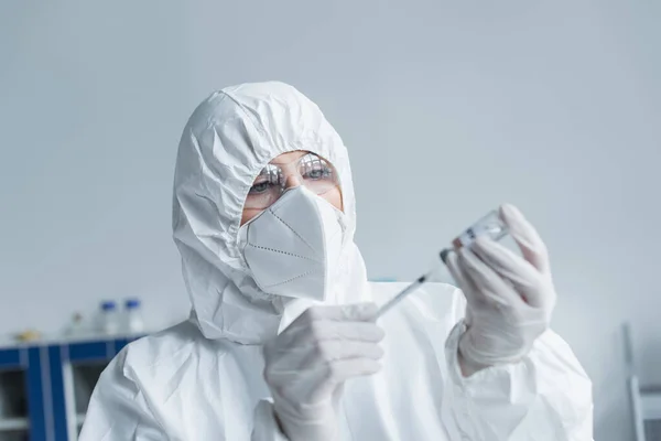 Ученый в защитном костюме и очках держит шприц и вакцину в лаборатории — стоковое фото