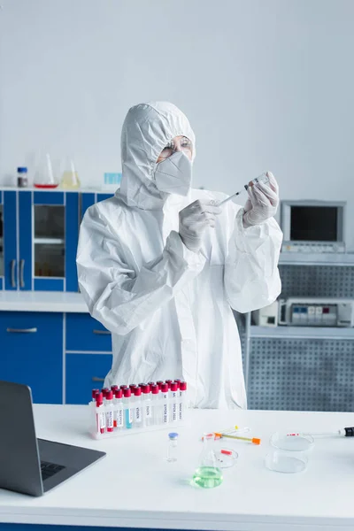Scientist in hazmat suit holding syringe and vaccine near test tubes in lab — Foto stock