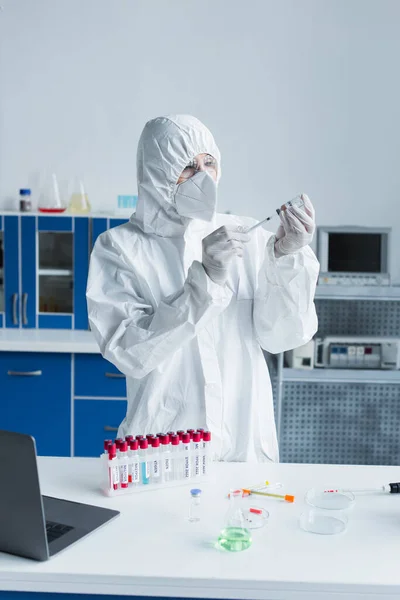 Scientist in hazmat suit holding syringe and vaccine near test tubes in lab — стоковое фото