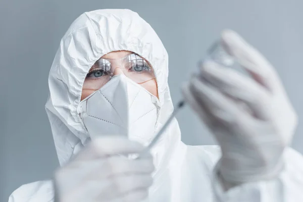 Scientist in hazmat suit and goggles holding blurred syringe and vaccine in lab — стоковое фото