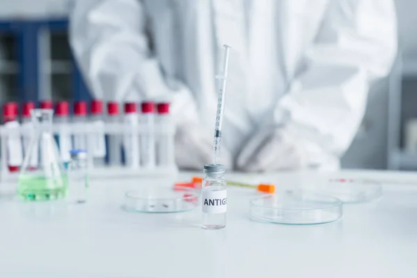 Cropped view of syringe in antigen near blurred petri dishes and scientist in lab — Stock Photo