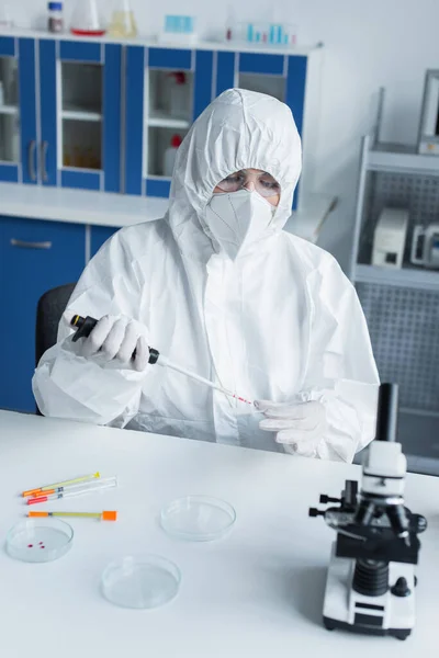 Cientista em luvas de látex e terno hazmat segurando pipeta eletrônica e placa de Petri em laboratório — Fotografia de Stock
