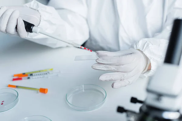 Cropped view of scientist in hazmat suit holding glass and electronic pipette in lab - foto de stock