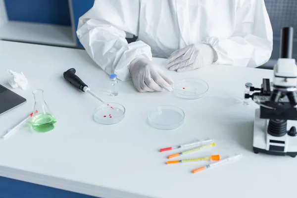 Cropped view of scientist in hazmat suit holding glass near petri dishes and microscope in lab — Stock Photo