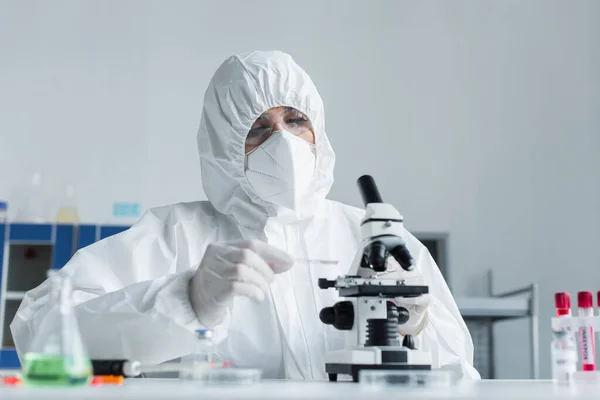 Wissenschaftler im Schutzanzug hält Glas, während er im Labor mit dem Mikroskop arbeitet — Stockfoto