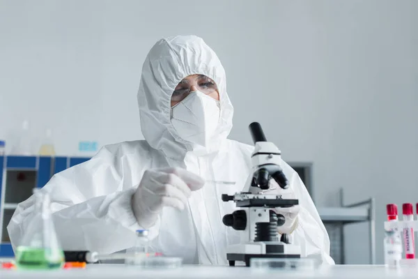 Scientist in hazmat suit holding glass while working with microscope in laboratory — стоковое фото