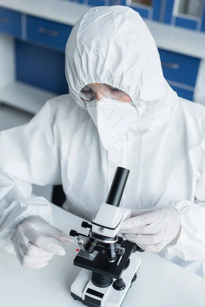 Scientist in hazmat suit holding glass near microscope in lab — Stockfoto