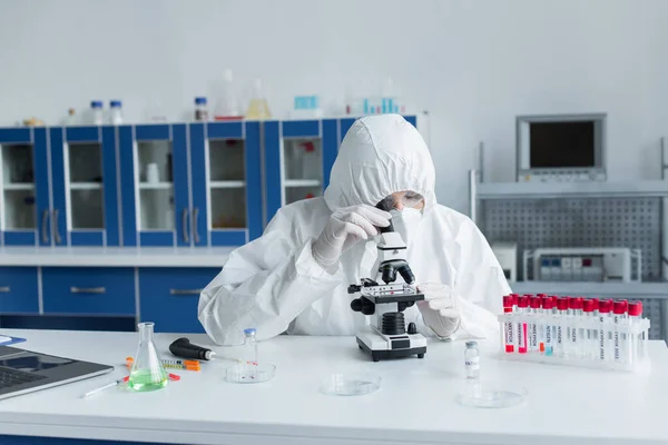 Ученый в защитном костюме с помощью микроскопа возле пробирки с надписью 