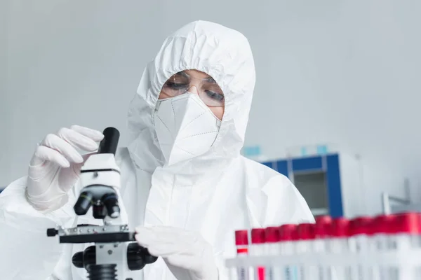 Scientist in hazmat suit using microscope near blurred test tubes in lab — Stock Photo