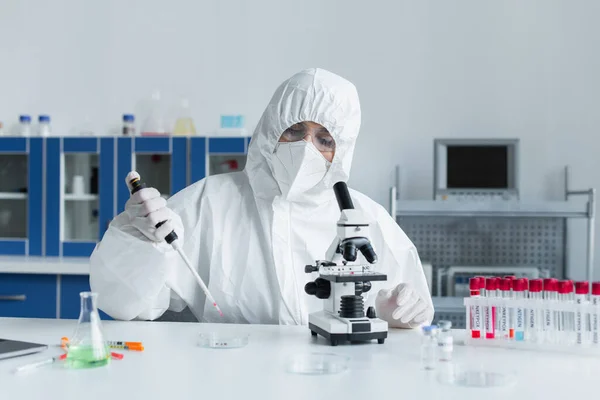 Wissenschaftler im Schutzanzug hält elektronische Pipette in der Nähe von Mikroskop und Reagenzglas im Labor — Stockfoto