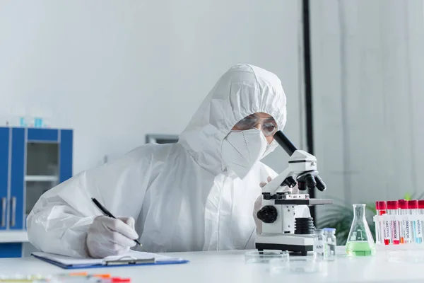 Scientist in hazmat suit looking in microscope near clipboard and test tubes in lab — Stock Photo