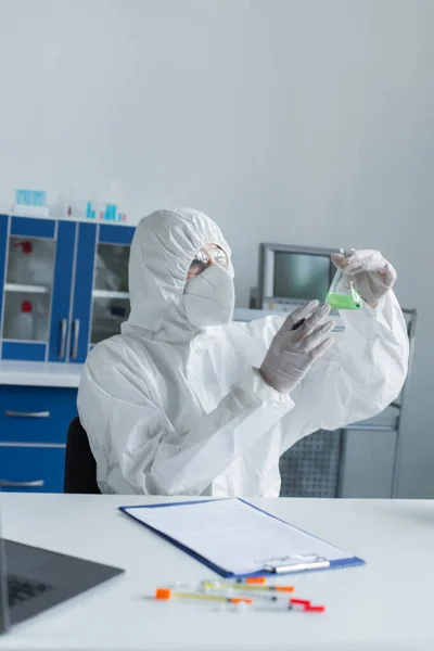 Scientist in hazmat suit holding flask near blurred clipboard and laptop in laboratory — Fotografia de Stock