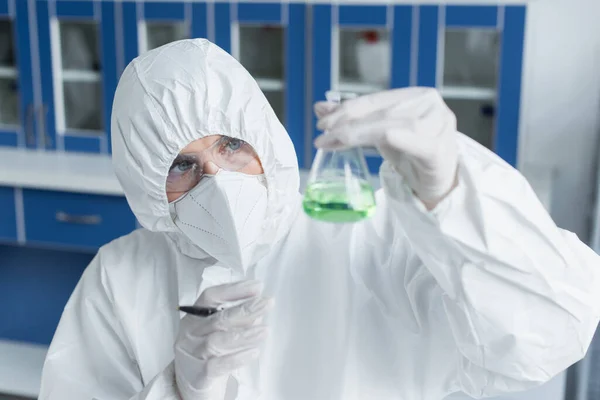 Scientist in protective suit and mask holding flask with liquid in lab — Stock Photo