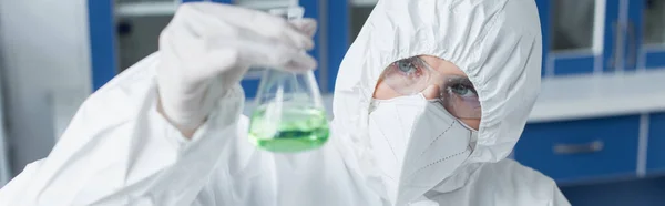 Scientist in hazmat suit and protective mask holding flask with liquid in lab, banner — Stock Photo