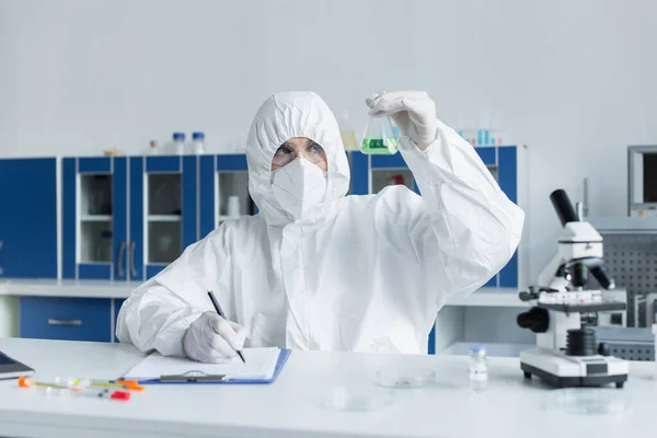 Cientista em terno hazmat trabalhando com frasco e escrevendo na área de transferência em laboratório — Fotografia de Stock