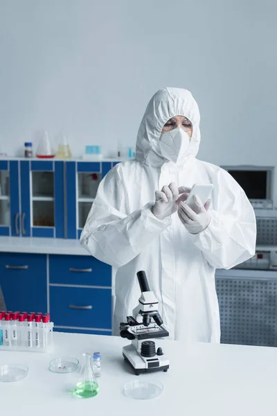 Scientist in hazmat suit using smartphone near microscope and test tubes in lab — Stock Photo