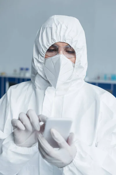 Scientist in hazmat suit and protective mask using blurred smartphone in lab — Photo de stock