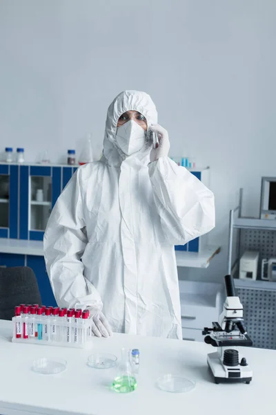 Вчений в захисному костюмі і масці розмовляє на смартфоні поблизу пробних трубок і мікроскопа в лабораторії — стокове фото