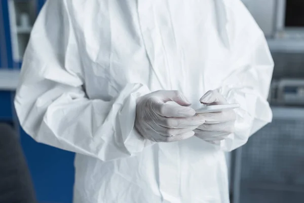 Cropped view of scientist in protective suit and latex gloves using mobile phone in lab — Fotografia de Stock